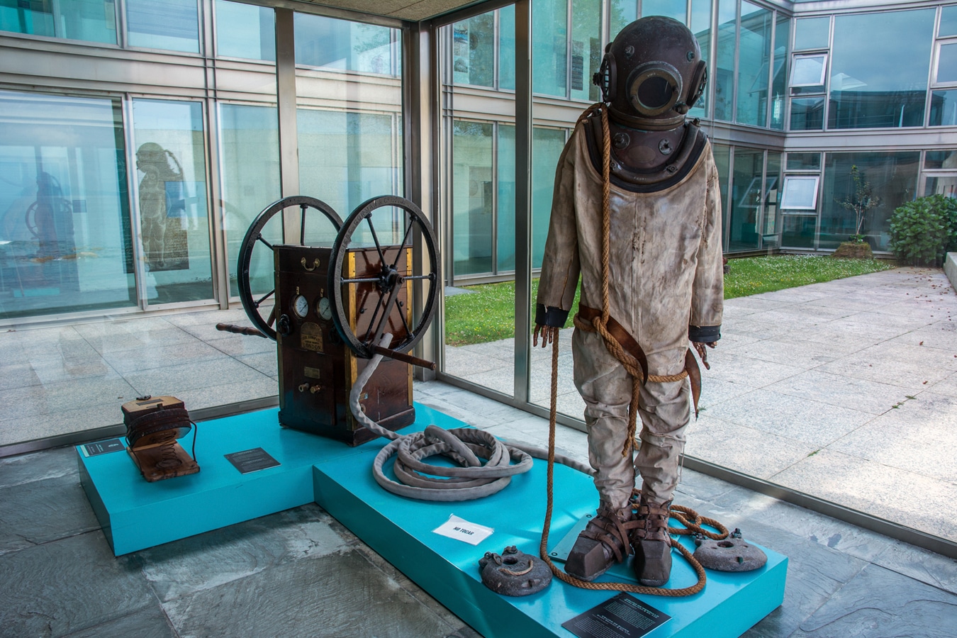 Ukázka potápěčského vybavení z první třetiny 20. stol., vzadu stojí dvouválcová vzduchová pumpa s ručním pohonem navržená pro zásobování vzduchem dvou potápěčů současně v střední hloubce nebo jednoho potápěče ve velké hloubce v expozici Muzea galicijského moře ve Vigu